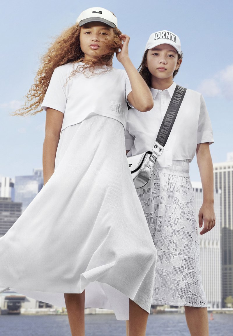 Deux mannequins portent des tenues blanches ; l'un est vêtu d'un chemisier court et d'une robe fluide, l'autre porte une chemise et une jupe à motifs. Elles portent toutes deux des casquettes.