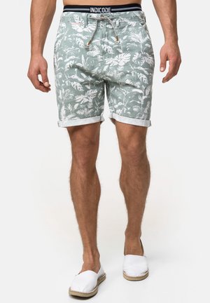 Shorts vert clair avec des motifs de feuilles tropicales blanches, aux revers roulés et à taille avec cordon de serrage. Portés avec des chaussures blanches à enfiler.