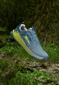 Μπλε και κίτρινο παπούτσι τρεξίματος σε μονοπάτι, αιωρούμενο πάνω από μια δασική επιφάνεια με χαλίκια και χόρτο, με φόντο πράσινα φυλλώματα.