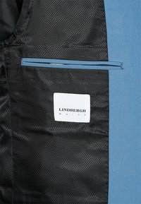 Interior de un blazer azul con un forro de malla negra, que presenta un bolsillo con ribete azul claro y una etiqueta blanca que dice "LINDBURGH WHITE."
