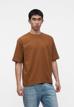 Jeune homme aux cheveux courts noirs portant une chemise marron ample à manches courtes et un jean bleu clair sur un fond uni.