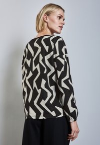 Haut à manches longues noir et beige présentant un motif abstrait en zigzag. Tissu doux et léger avec une coupe décontractée et des poignets élastiques.