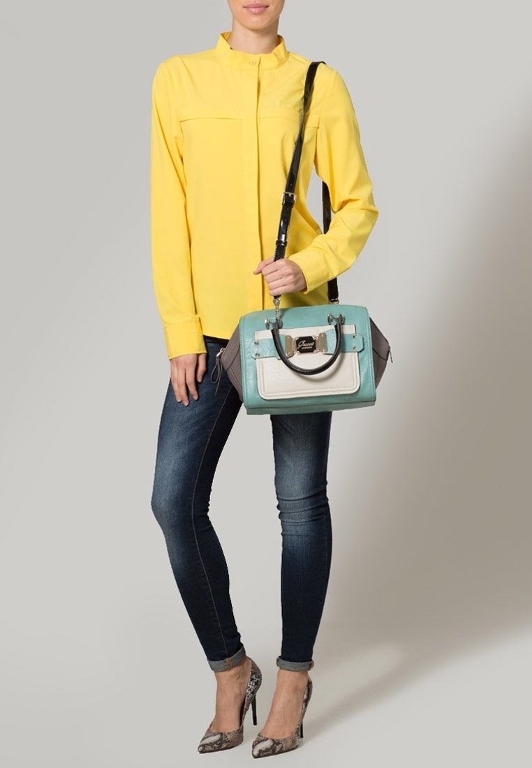 Chemise jaune à manches longues, jean skinny noir et escarpins à imprimé serpent gris. Tient un sac à main multicolore avec un design turquoise et beige.
