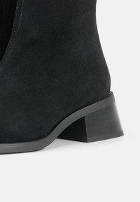 Svart semsket ankelstøvel med rund tå, middels høy stablet hæl og glidelås på siden. Glatt tekstur, slankt design.