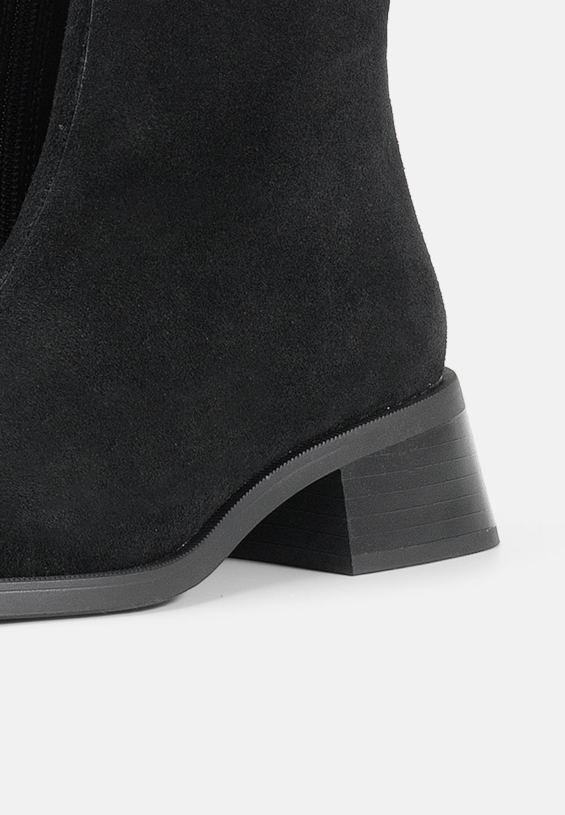 Bottine en suède noir avec un bout arrondi, un talon empilé de hauteur intermédiaire et une fermeture éclair sur le côté. Texture lisse, design épuré.