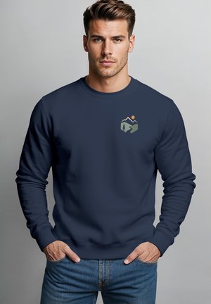 Mann mit kurzen braunen Haaren trägt einen marineblauen Pullover mit einem kleinen Natur-Logo mit Bergen, einem Baum und einer Sonne auf der Brust.