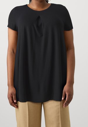 Femme portant un haut noir ample à manches courtes avec un petit pli à l'avant, associé à un pantalon beige à jambes larges sur un fond uni.