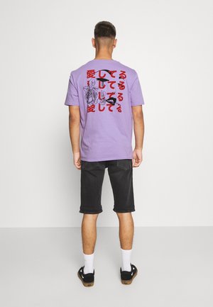 Homme debout de dos portant un T-shirt violet avec des caractères japonais rouges et des motifs floraux et de visage noirs, short noir, chaussettes blanches, chaussures noires.
