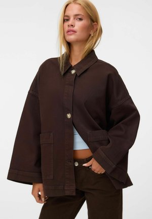 Femme blonde portant une veste oversize marron foncé avec de gros boutons et poches, un haut bleu clair et un pantalon marron foncé assorti.
