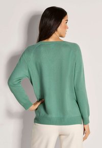 Frau trägt einen langärmeligen, mintgrünen Pullover und weiße Hosen, steht mit einer Hand auf der Hüfte und blickt von der Kamera weg.