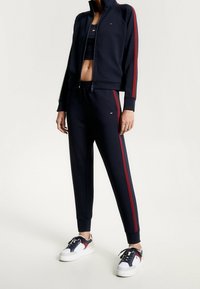 Giacca navy con zip e strisce rosse, abbinata a pantaloni jogger coordinati e scarpe bianche con dettagli rossi e grigi. Tessuto morbido, design aderente.