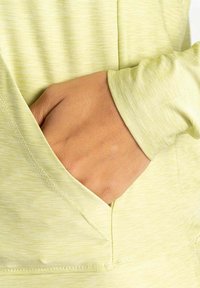 Hellgrünes sportliches Hoodie mit strukturierter Oberfläche, ausgestattet mit einer Kängurutasche und engen Bündchen, die die Dehnbarkeit und Atmungsaktivität des Stoffes betont.