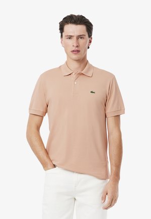Ljus persikofärgad polo med krage, korta ärmar och en broderad grön krokodil-logotyp. Bärs med vita byxor, har en mjuk textur.