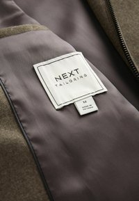 Die Innenseite der braunen Jacke zeigt ein glattes, graues Futter und ein weißes Etikett mit "NEXT TAILORING" und der Größe "M", auf dem steht "HERGESTELLT IN VIETNAM."