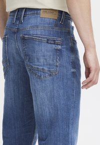 Un jeans en denim bleu porté par une personne, montrant une poche arrière avec une petite étiquette noire et une étiquette de marque en cuir marron sur la taille.