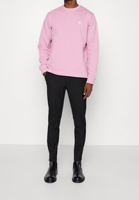 Felpa rosa a maniche lunghe con un piccolo logo dorato, abbinata a pantaloni neri a sigaretta e stivaletti neri, in piedi di fronte a uno sfondo neutro.