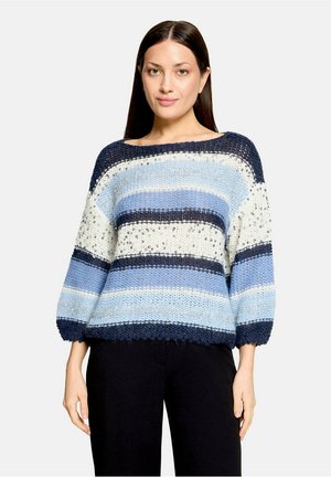 Femme aux cheveux raides et foncés portant un pull tricoté ample avec des rayures horizontales bleues, blanches et bleu marine et un pantalon noir, debout et face à l'avant.