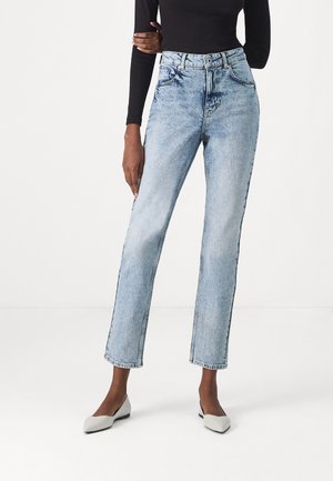 Femme portant un jean bleu clair taille haute à jambes droites, un top noir à manches longues, et des ballerines blanches à bout pointu, debout avec un bras croisé.