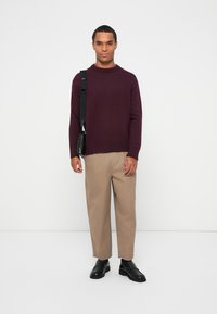 Μαρό μανίκι πλεκτό πουλόβερ, μπεζ παντελόνι, μαύρα παπούτσια. Το μοντέλο κρατά μια μικρή μαύρη τσάντα με ιμάντα ώμου. Απλός, μοντέρνος σχεδιασμός, λεία υφή.