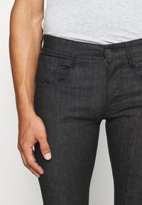 Jeans de mezclilla negros con un ajuste slim, diseño de cinco bolsillos y cierre de botón en la parte frontal. La tela presenta una ligera textura con un acabado suave.