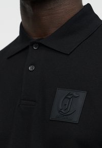 Μαύρο πόλο πουκάμισο με δύο κουμπιά και κεντημένο λογότυπο σε ύφασμα που γράφει "Just Cavalli" στο αριστερό μέρος του στήθους, φορεμένο από άτομο με σκουρόχρωμο δέρμα.