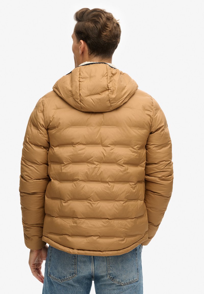 Bruine gewatteerde jas met een capuchon, met een gestikt ontwerp en een gladde textuur. Draagt over lichtblauwe jeans, gezien vanaf de achterkant.