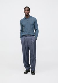 Polo azul de manga comprida com gola e três botões, combinado com calças plissadas cinza claro e sapatos pretos.