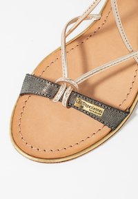 Sandalo con cinturini beige e fascia nera testurizzata, dotato di una placca con logo dorato e una soletta in pelle marrone con dettagli di cucitura.