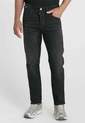 Uomo che indossa jeans neri e scarpe da ginnastica grigie, in piedi con una mano in tasca su uno sfondo semplice.