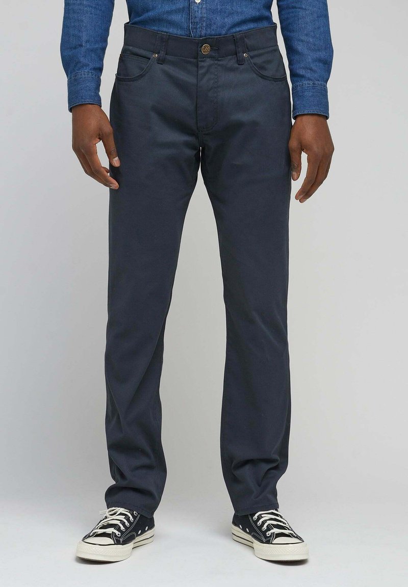 Marineblaue Baumwollhose mit geradem Schnitt, ausgestattet mit vorderen Taschen, einem Knopfverschluss und getragen mit schwarzen Sneakers im Punktedesign.