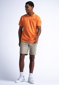 Oranje katoenen polo shirt met een kraag en logo-detail, gecombineerd met beige shorts en witte sneakers. De outfit heeft een casual, ontspannen pasvorm.