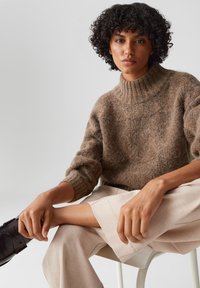 Maglione marrone a maglia grossa con colletto alto a costine, indossato su pantaloni leggeri e dal taglio morbido. Texture liscia e silhouette rilassata.