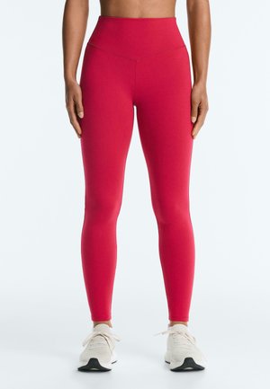 Kvinde iført højttaljede lyserøde leggings og hvide sneakers, stående med arme afslappet langs siderne på en ensfarvet hvid baggrund.