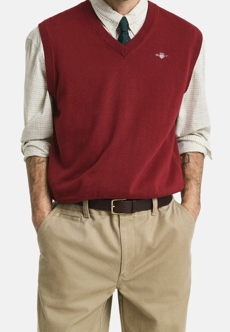 Homme portant un gilet rouge à col en V par-dessus une chemise à carreaux aux manches relevées, pantalon beige et cravate vert foncé, mains dans les poches.