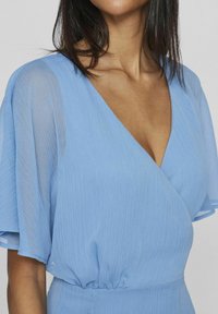 Svetlo modré šaty s výstrihom do V, skladanou textúrou a volnými krátkymi rukávmi. Pás je jemne zhabaný pre prispôsobený vzhľad.