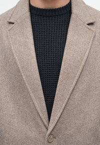 Sacou dintr-un amestec de lână bej, cu rever clasic, închidere cu un singur nasture și un pulover texturat de tricot de culoare bleu închis sub el.