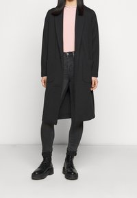 Largo abrigo negro con frente abierto, dos bolsillos frontales, llevado sobre una blusa rosa claro, combinado con jeans ajustados oscuros y botas negras de cordones.