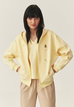 Žena s čiernymi vlasmi, oblečená v bledozltej mikine na zips s vyšívanými kvetmi a včelami, so zodpovedajúcim žltým tričkom a béžovými nohavicami.