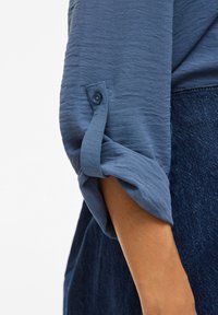 Blaues, strukturiertes Hemd mit hochgekrempeltem Ärmel, der mit einem Knopf befestigt ist. Der Ärmel und der Arm sind vor einem dunklen Denim-Hintergrund zu sehen.