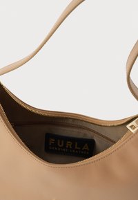Beżowa skórzana torebka o gładkiej fakturze, z złotym zamkiem błyskawicznym i czarną wewnętrzną metką z napisem „FURLA GENUINE LEATHER”.