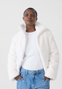 Witte fleecejack met ritssluiting en hoge kraag, zachte textuur en een relaxte pasvorm, gedragen over een wit shirt en in combinatie met lichtblauwe denim jeans.