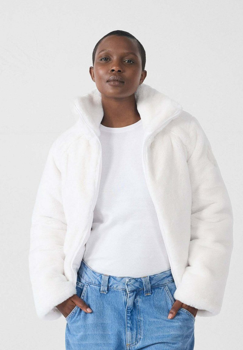 Witte fleecejack met ritssluiting en hoge kraag, zachte textuur en een relaxte pasvorm, gedragen over een wit shirt en in combinatie met lichtblauwe denim jeans.