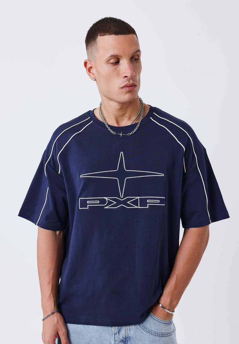 Homme portant un T-shirt oversize bleu marine avec des liserés blancs et le logo "PXP", assorti à un jean bleu clair et un collier chaîne argenté.