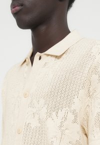 Μπεζ πλεκτό πόλο πουκάμισο με ανάγλυφο σχέδιο, κλείσιμο με κουμπιά, γιακά και ανοιχτό πλέγμα, προσφέροντας έναν ελαφρύ σχεδιασμό.