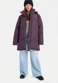 Γυναίκα φορώντας μαύρο πλεκτό σκουφάκι, μωβ καπιτονέ μπουφάν, λιλά πουλόβερ, ανοιχτόχρωμα παντελόνια με φαρδιά πόδια και μαύρα παπούτσια, σταθμευμένη σε λευκό φόντο.