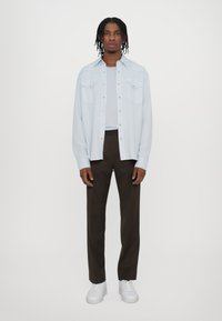 Svetlo modrá košeľa s gombíkmi a dvoma prsnými vreckami, skombinovaná s bielym tričkom a tmavohnedými nohavicami. Biely tenisky dotvárajú celý outfit.