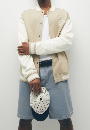 Person iført beige og hvid varsityjakke, hvid skjorte, grå shorts, holdende en beige og marineblå stribet kasket foran en ensfarvet baggrund.