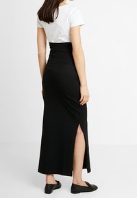 Femme portant une jupe maxi noire taille haute avec une fente sur le côté, un haut blanc à manches courtes et des mocassins noirs, se tenant devant un fond blanc.