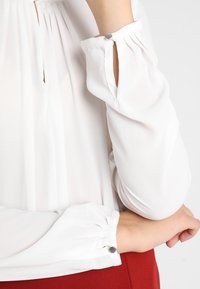 Witte blouse met lange,Gathered mouwen en kleine zilveren knoopaccenten bij de manchetten. De stof lijkt lichtgewicht en iets doorschijnend.