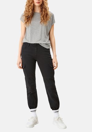 Frau trägt ein graues Kurzarmshirt, schwarze Cargohosen, weiße Socken mit Streifen und weiße Turnschuhe und steht vor einem schlichten Hintergrund.